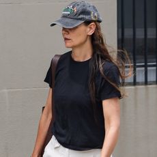 Katie Holmes walking in New York City wearing a baseball cap, black T-shirt, white trousers, and almond-toe black flats.