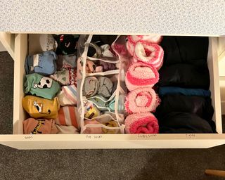 Open and organized sock drawer showing left to right colorful sock bundles with cartoon cats, stripes and Christmas trees, a mesh organizer with rows of colorful ankle sock bundles, a row of fluffy pink socks and black tights folded into rows. There are correlating labels on the front top edge of the drawer