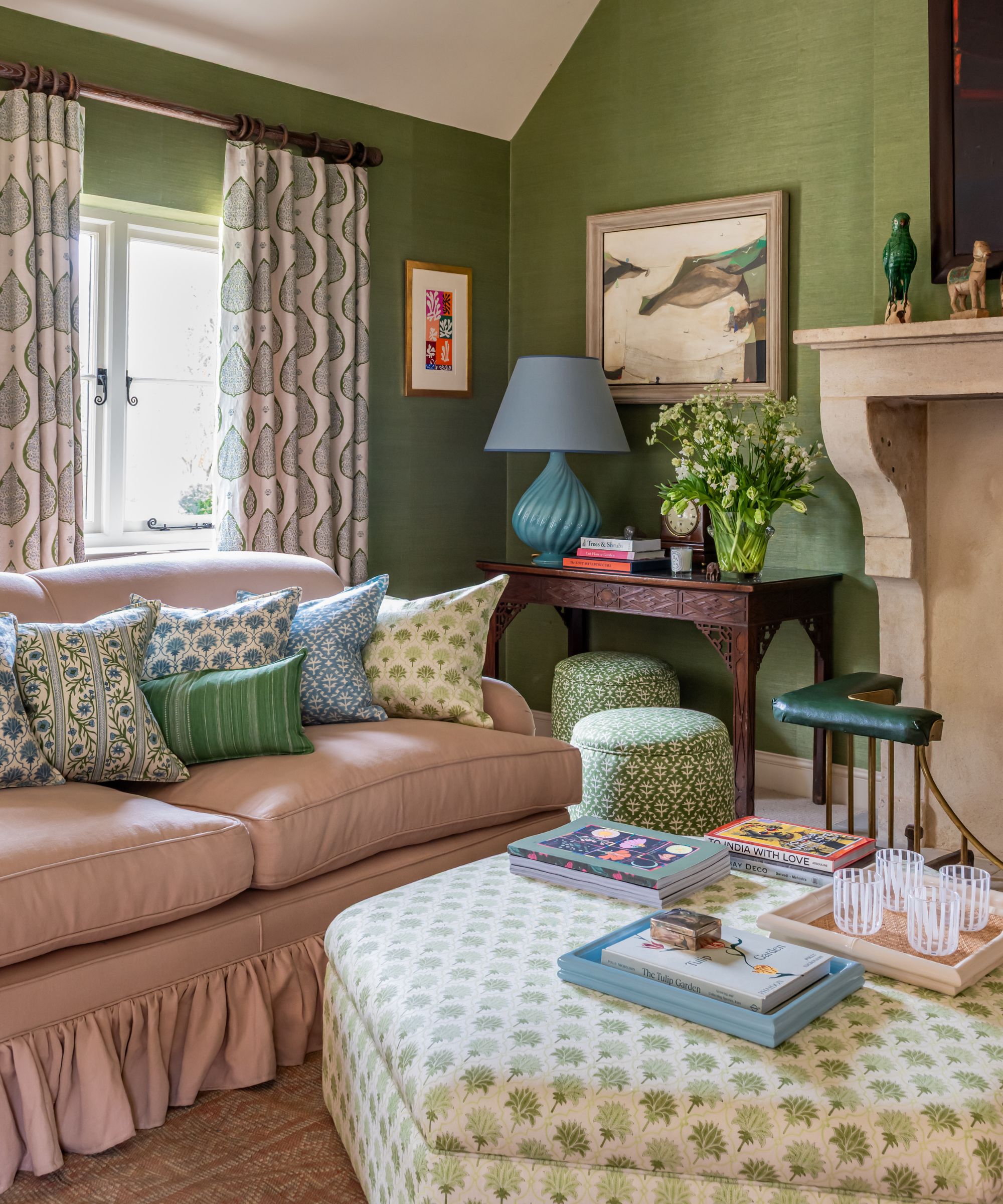 A well-lit interior featuring green grasscloth walls and a cream stone fireplace. A pale pink sofa with a ruffled base is layered with various floral and geometric cushions. In the foreground, a large hexagonal ottoman with a green leaf pattern holds books and a tray. Matching patterned curtains hang by a window, and two small green garden stools are tucked under a side table.