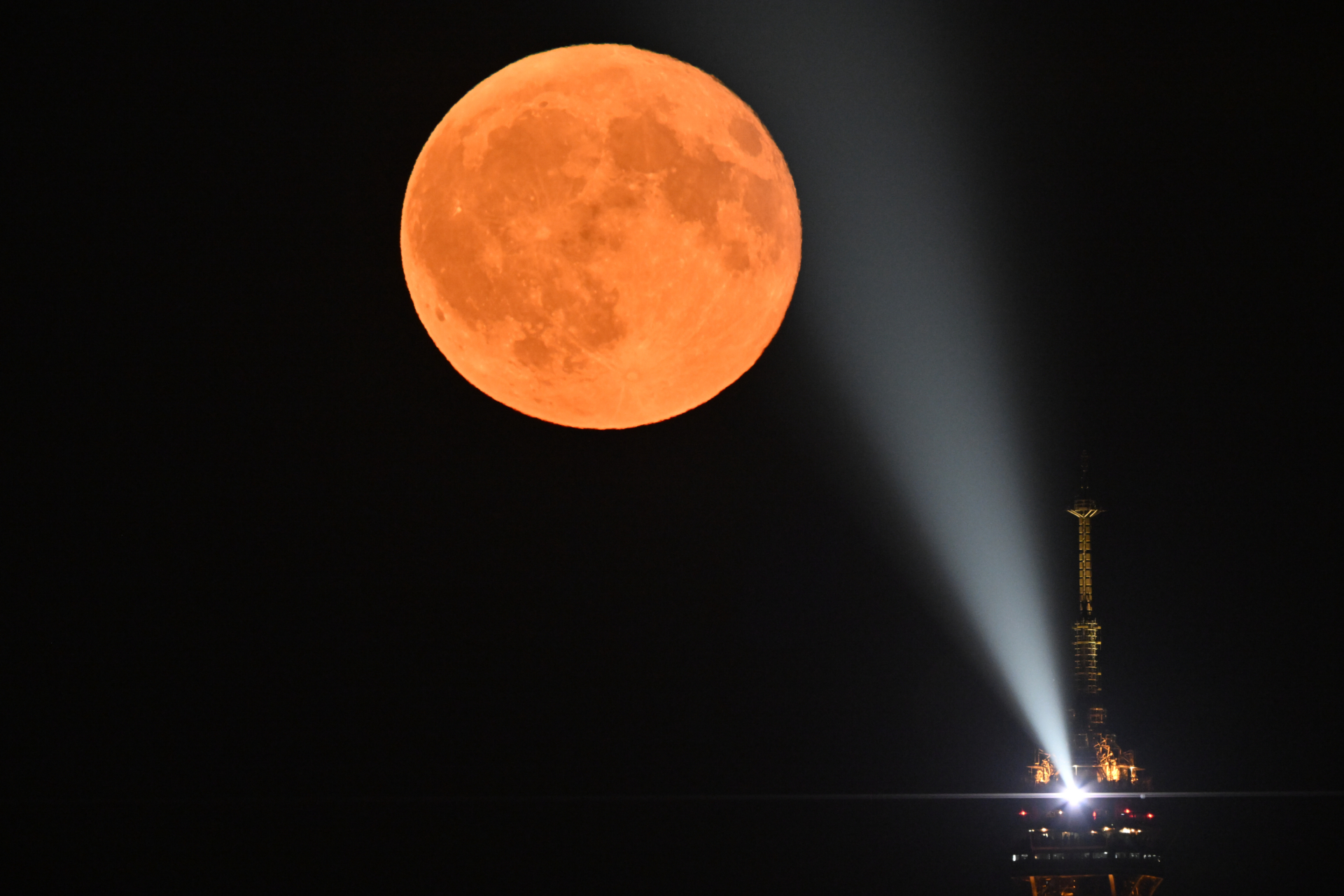 A spotlight is pictured shining on an orange full moon, emanating from the very tip of the Eiffel Tower in Paris, which is pictured to the lower right of the image against a black night sky.