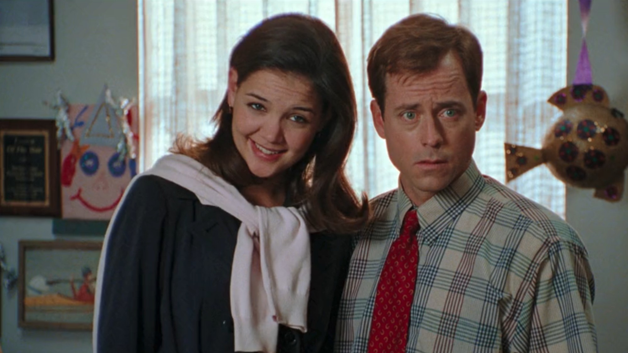 Katie Holmes looking skeptical, wearing a dark dress with a light-colored sweater tied around her neck. She is standing with Greg Kinnear, wearing a shirt and tie in The Gift.
