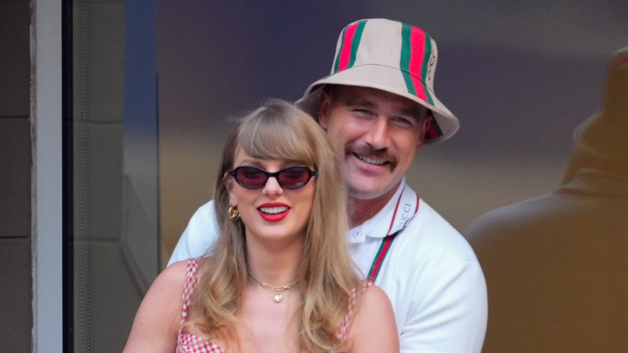 Taylor Swift wears sunglasses and bright red lipstick while Travis Kelce puts his arms around her while wearing a Gucci bucket hat