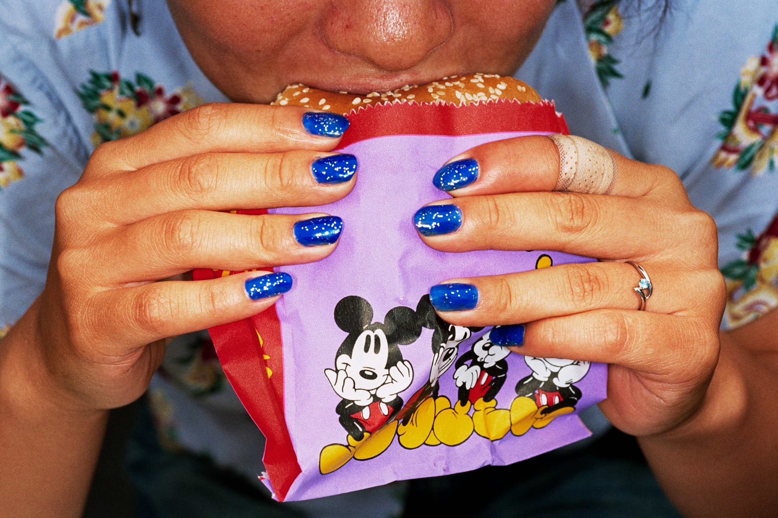 A close-up shot of a person with sparkly blue nail polish holding a sesame seed burger wrapped in a Mickey Mouse paper sleeve.
