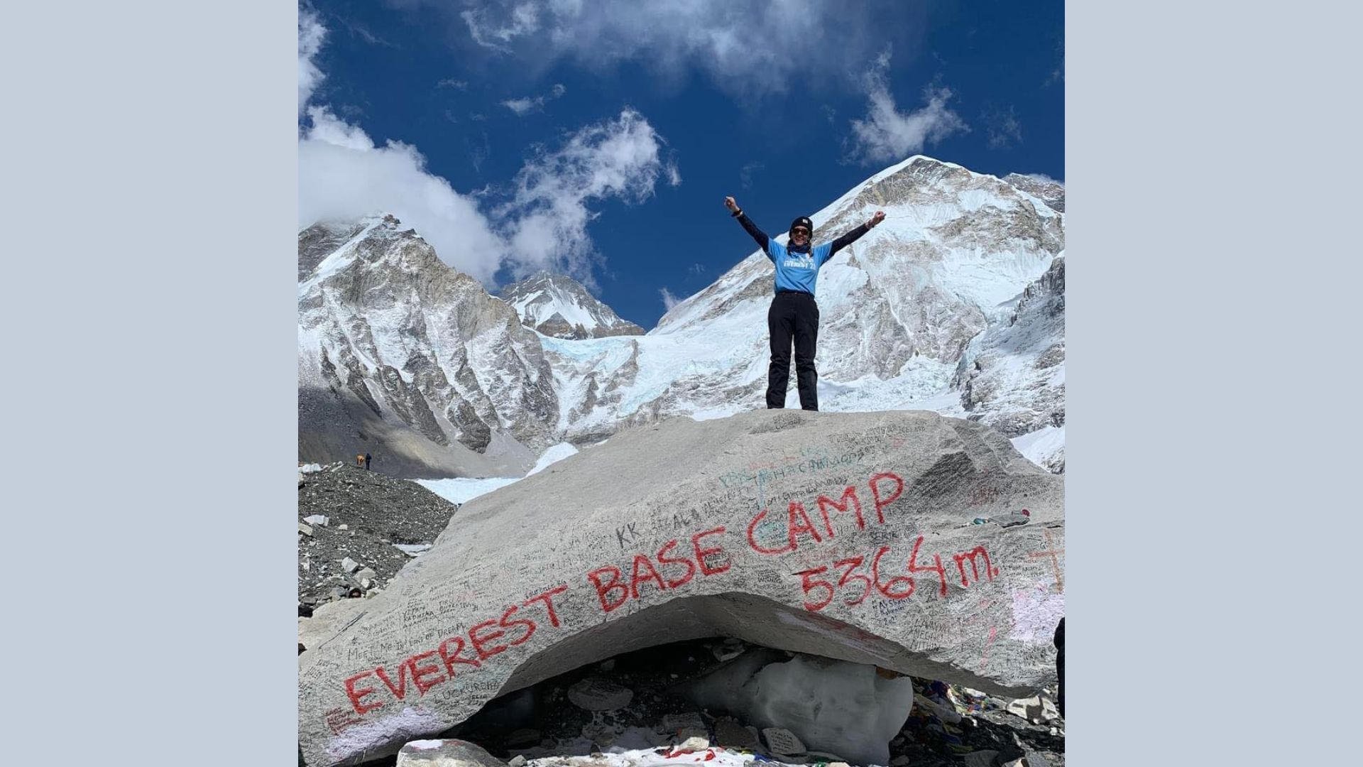 Stacey Kirkpatrick at Everest Base Camp, standing on rock with snowy mountains behind her