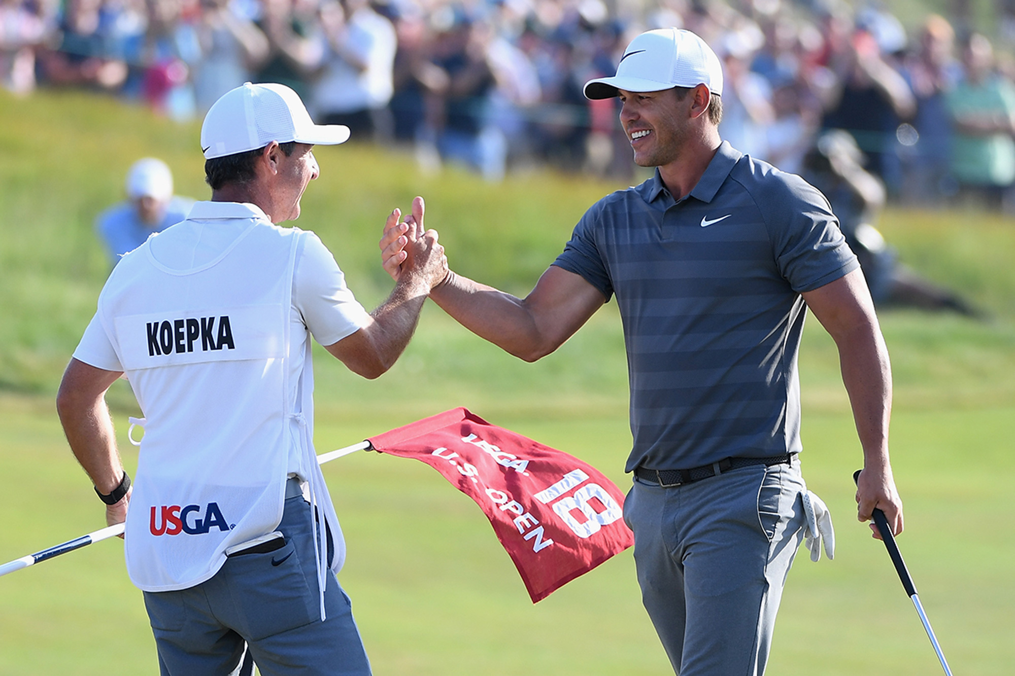 Brooks Koepka shaking hands with his caddie, who is holding the 18th flag, after winning the 2018 US Open at a score of +1, the last time the event was won at par or worse