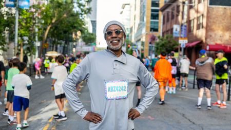 A vibrant, attractive older man poses at a race or competitive walk with a number pinned to his shirt. He wears sunglasses and a hat and smiles at the camera.