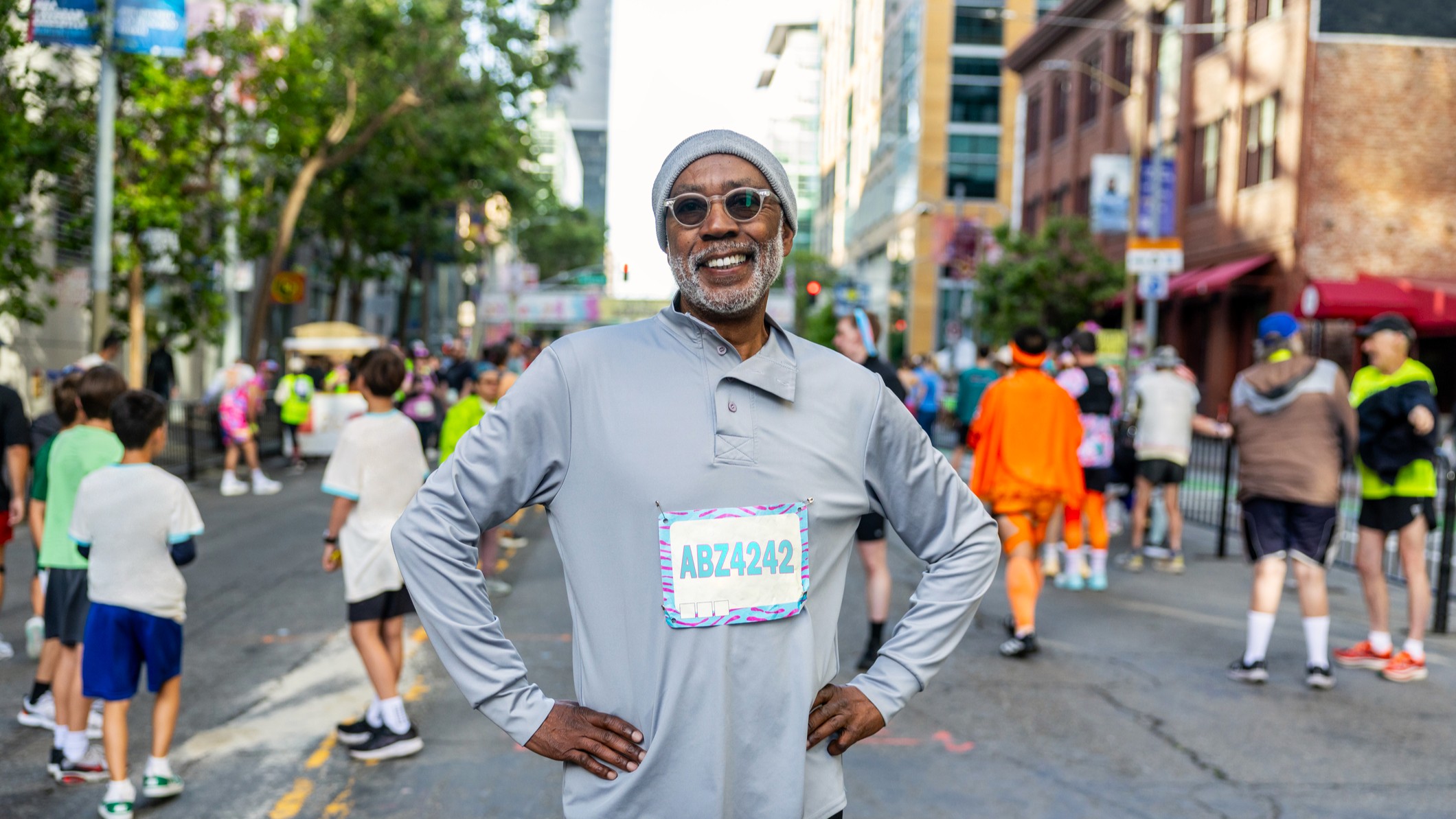 A vibrant, attractive older man poses at a race or competitive walk with a number pinned to his shirt. He wears sunglasses and a hat and smiles at the camera.