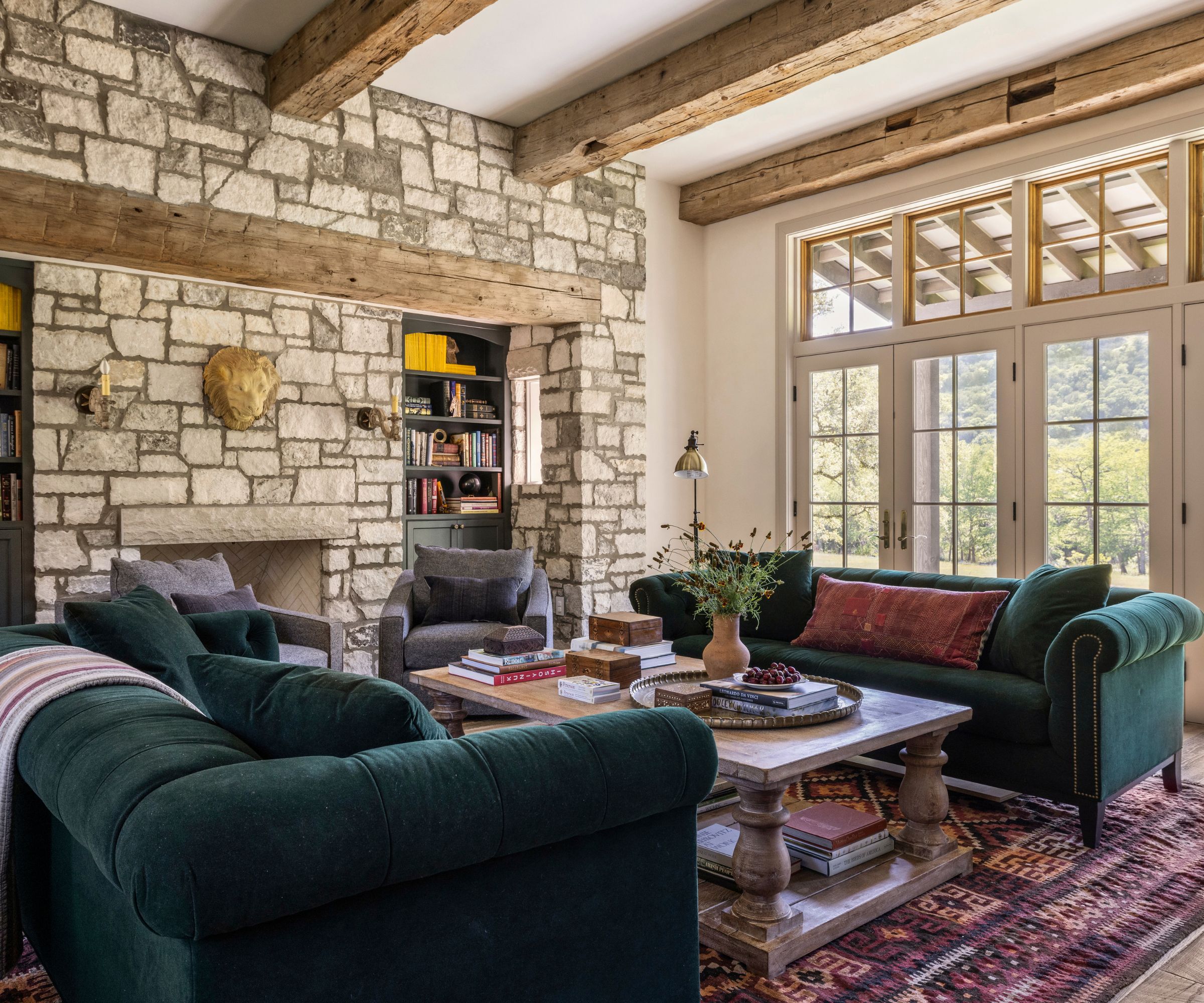 A rustic living room with a brick wall, wood ceiling beams, and wall of glass doors. Two emerald green sofas, a dark pink Persian rug, wood coffee table and eclectic decor.