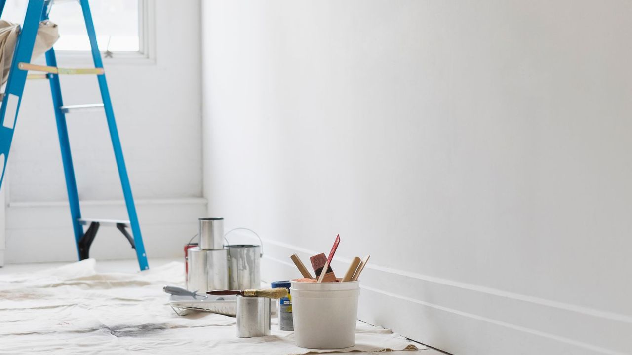 A blue step ladder and open tins of paint on a drop sheet in front of a blank wall