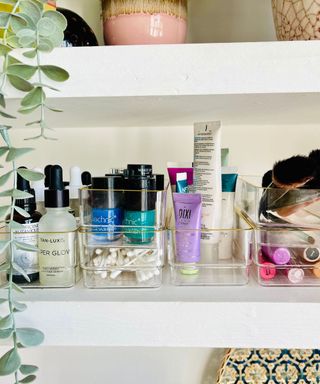 Clear plastic drawer organizers with a gold rim being used to organize small makeup items on white shelves