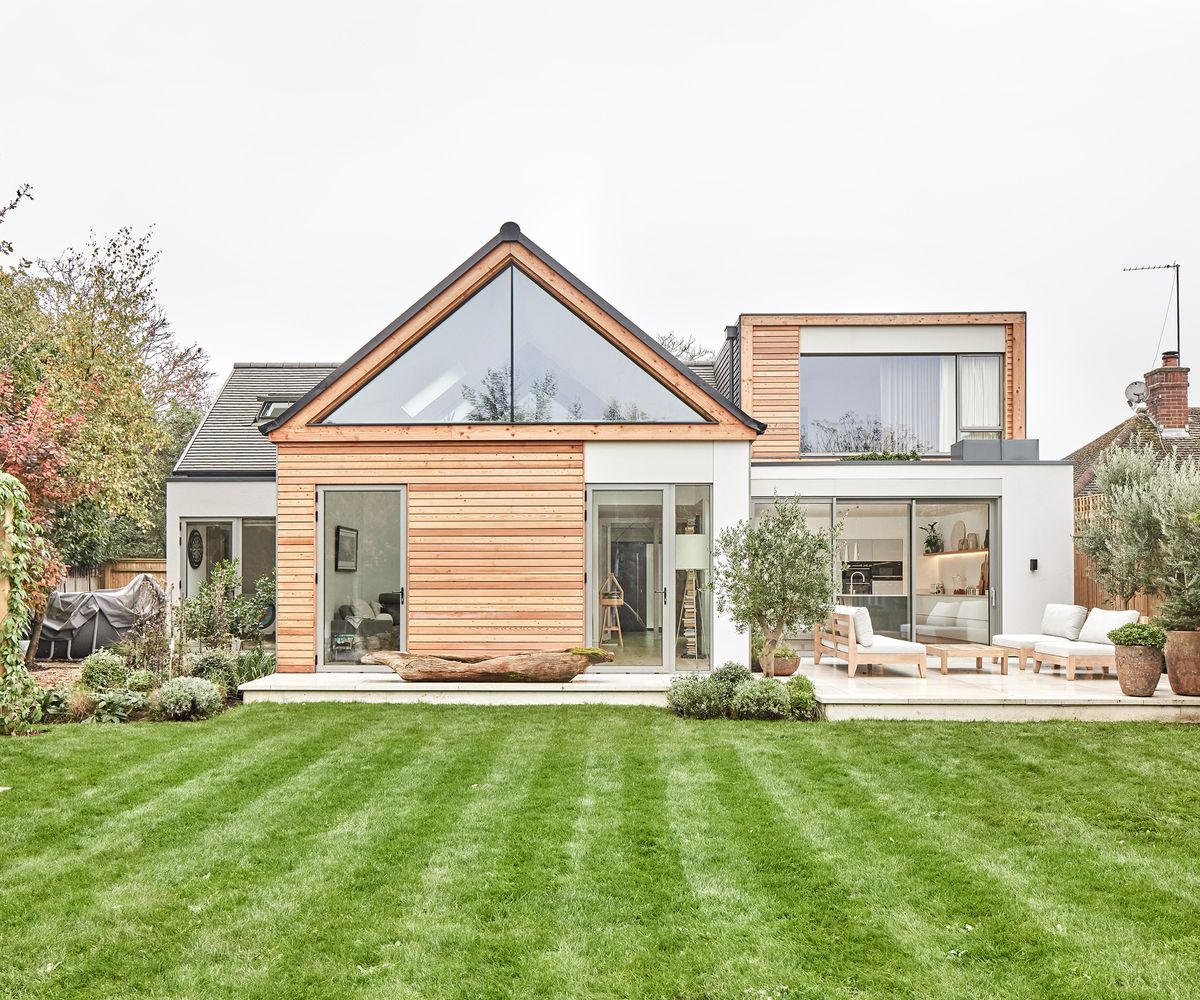 The rear exterior of a house with a lush green lawn and a contemporary self build home. The glazing is bespoke, and the cladding is timber