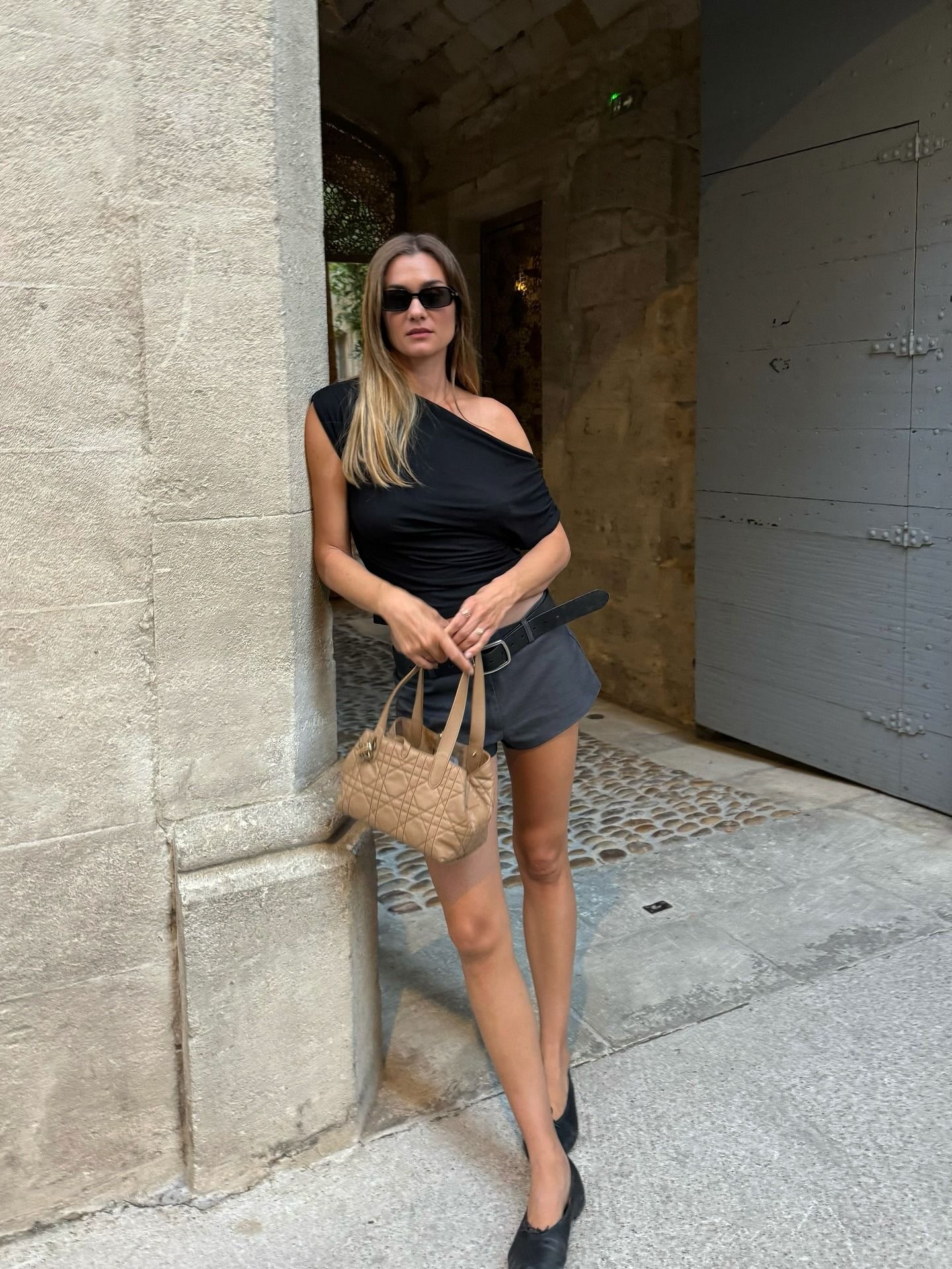 Influencer @annelauremais leans on a door frame wearing gray shorts, a black top, and black leather ballet flats.