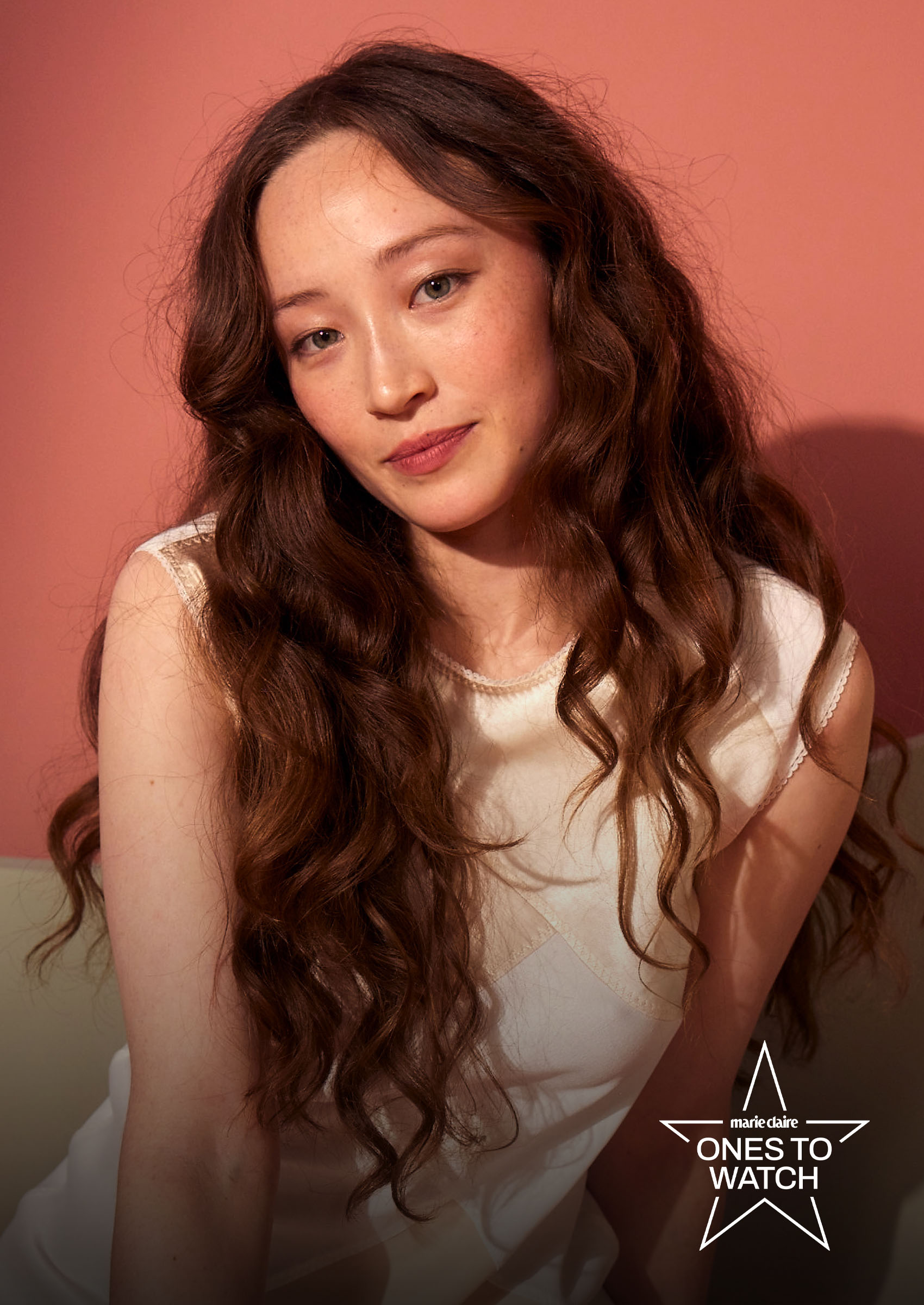 actress havana rose liu poses sitting down with her hair curly and in a white dress against a pink backdrop
