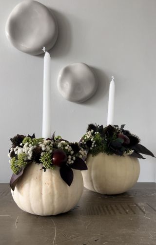 Gray wall and two white pumpkins on a table with flowers on them and white candle sticks