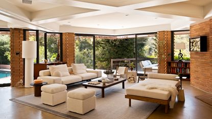 Image of an open concept living room that has windowed doors that open to the outdoors. There is a modern sofa, a modern chaise lounge, and two white ottomans around a wooden coffee table in the center of the room. 