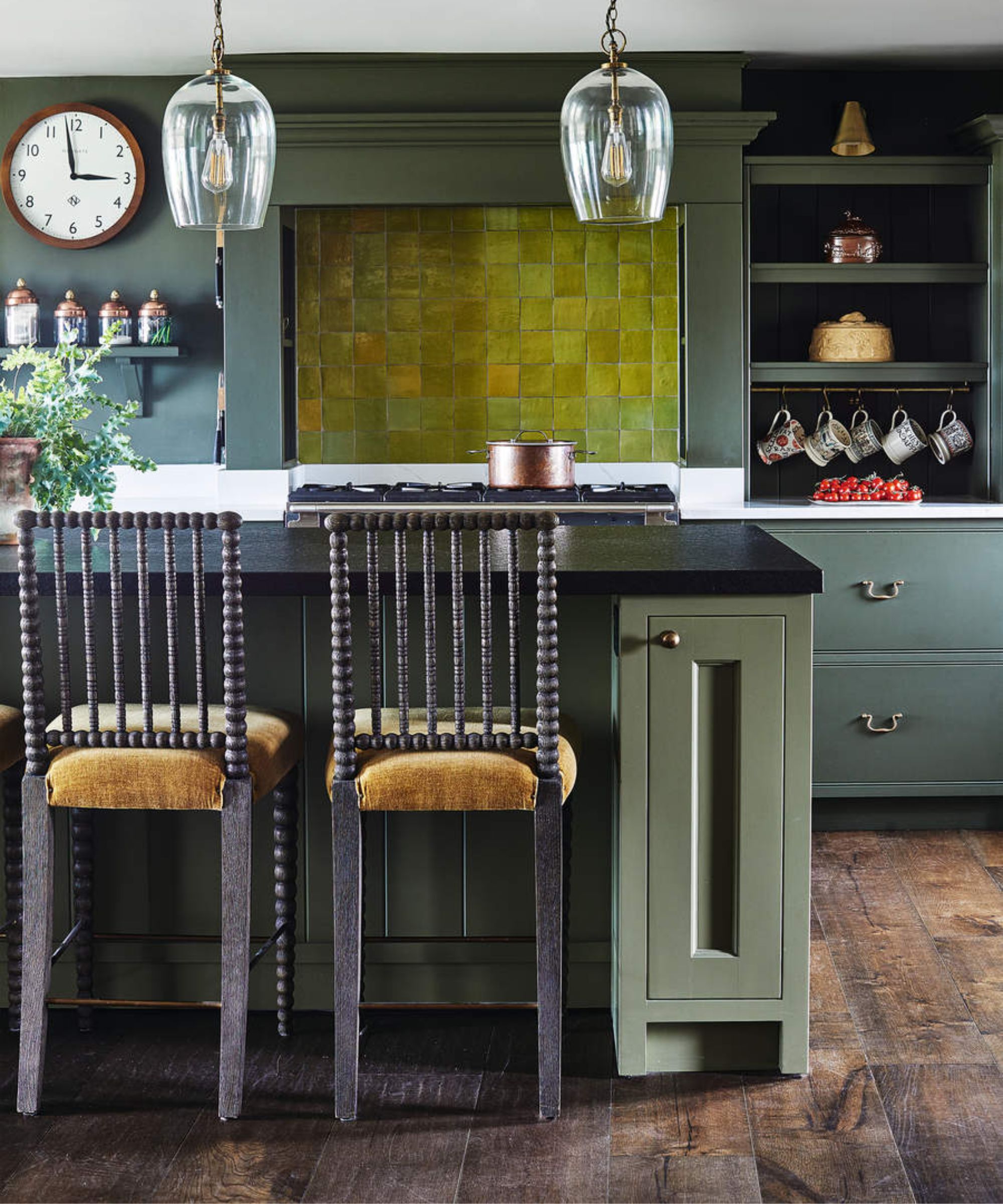A green kitchen with brown wooden flooring, green cabinets, green tiling, and a green kitchen island with a black marble countertop. In the foreground are two wooden chairs with velvet upholstery, with two glass pendant lamps hanging above. To the right is alcove shelving with mugs and to the left is a wooden circular wall clock.