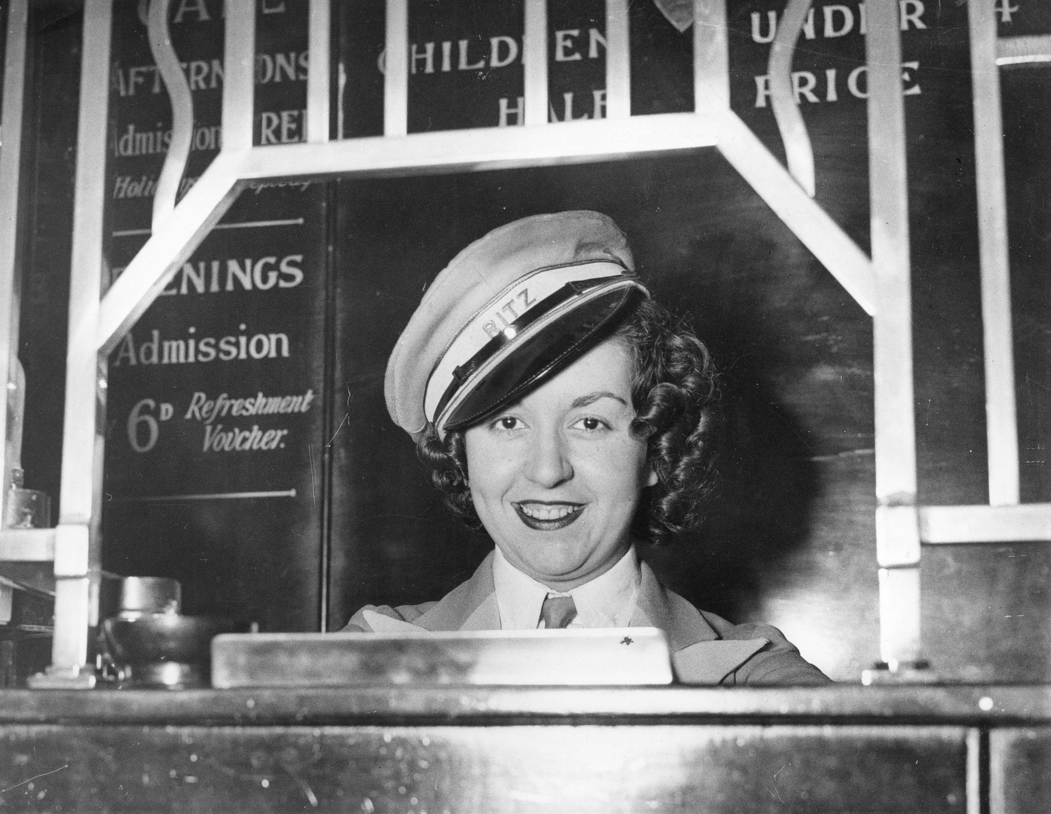 A smiling woman in a peaked cap bearing the word "Ritz" sits behind a dance hall pay desk, with signs behind her advertising admission prices and a 6d refreshment voucher.