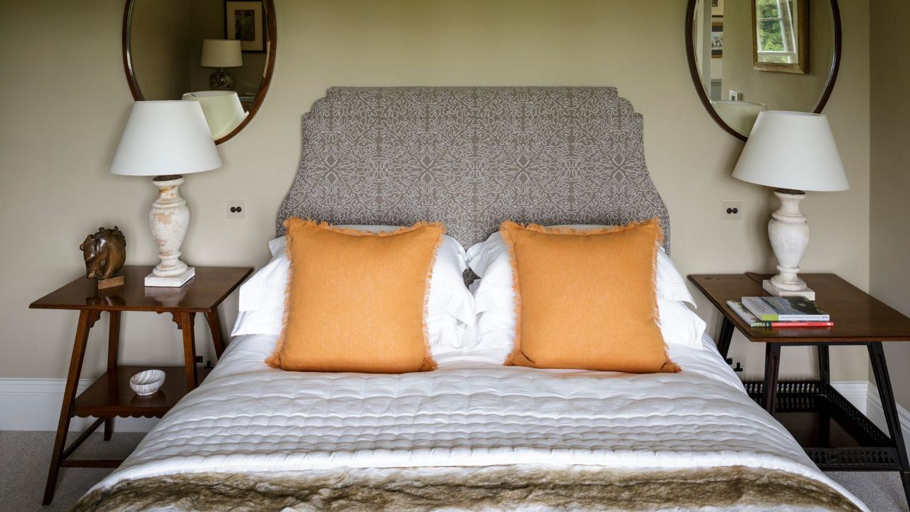 Cream bedroom with double bed with patterned light brown headboard, white bedding, and orange cushions. To either side of the bed are matching wooden bedside tables with white lamps and white lampshades, and above both are matching oval mirrors