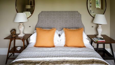 Cream bedroom with double bed with patterned light brown headboard, white bedding, and orange cushions. To either side of the bed are matching wooden bedside tables with white lamps and white lampshades, and above both are matching oval mirrors