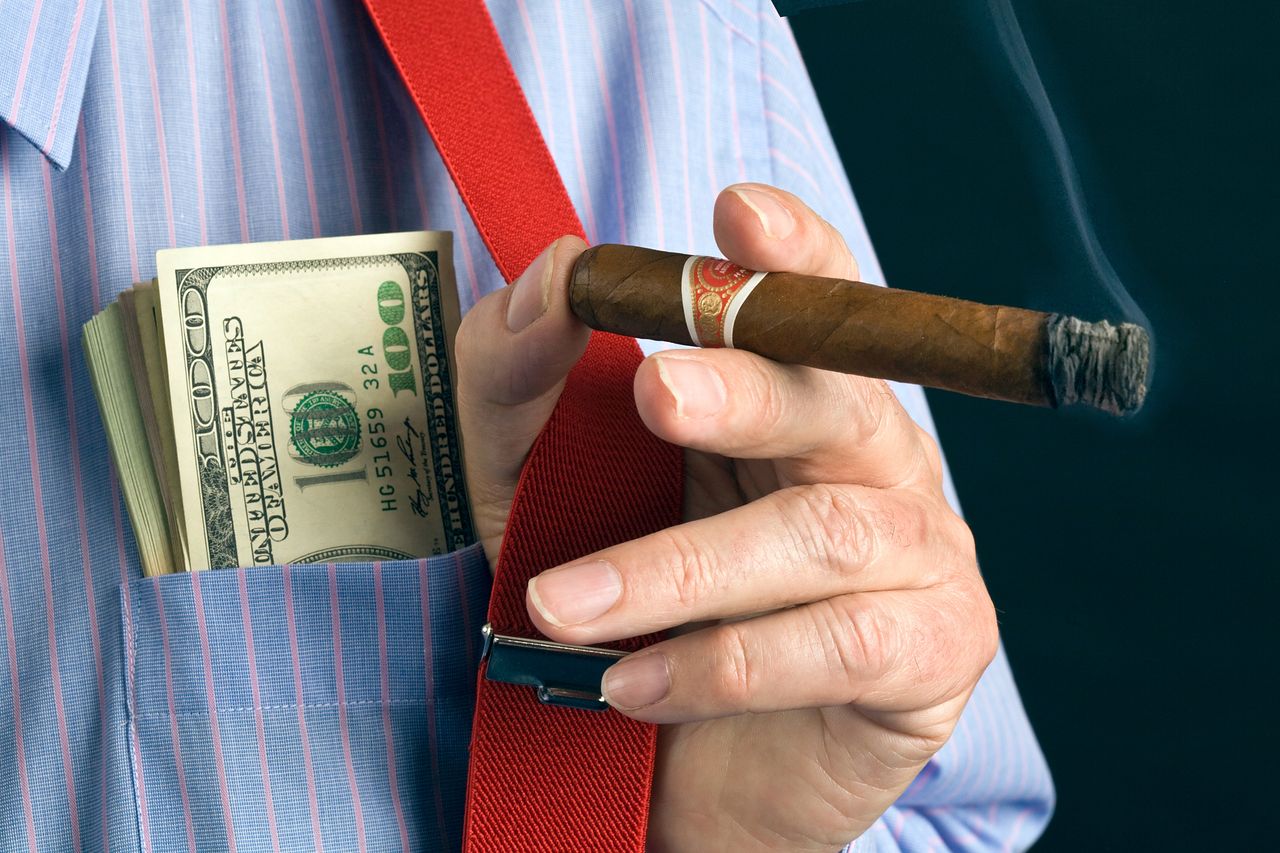 A close-up of a man&#039;s hand holding a cigar, with hundred dollar bills poking out of his breast pocket.