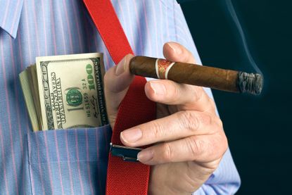 A close-up of a man's hand holding a cigar, with hundred dollar bills poking out of his breast pocket.