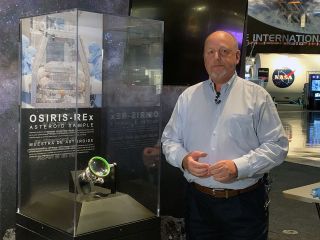a bald man in a light-blue shirt stands in front of a glass display case holding a piece of an asteroid.