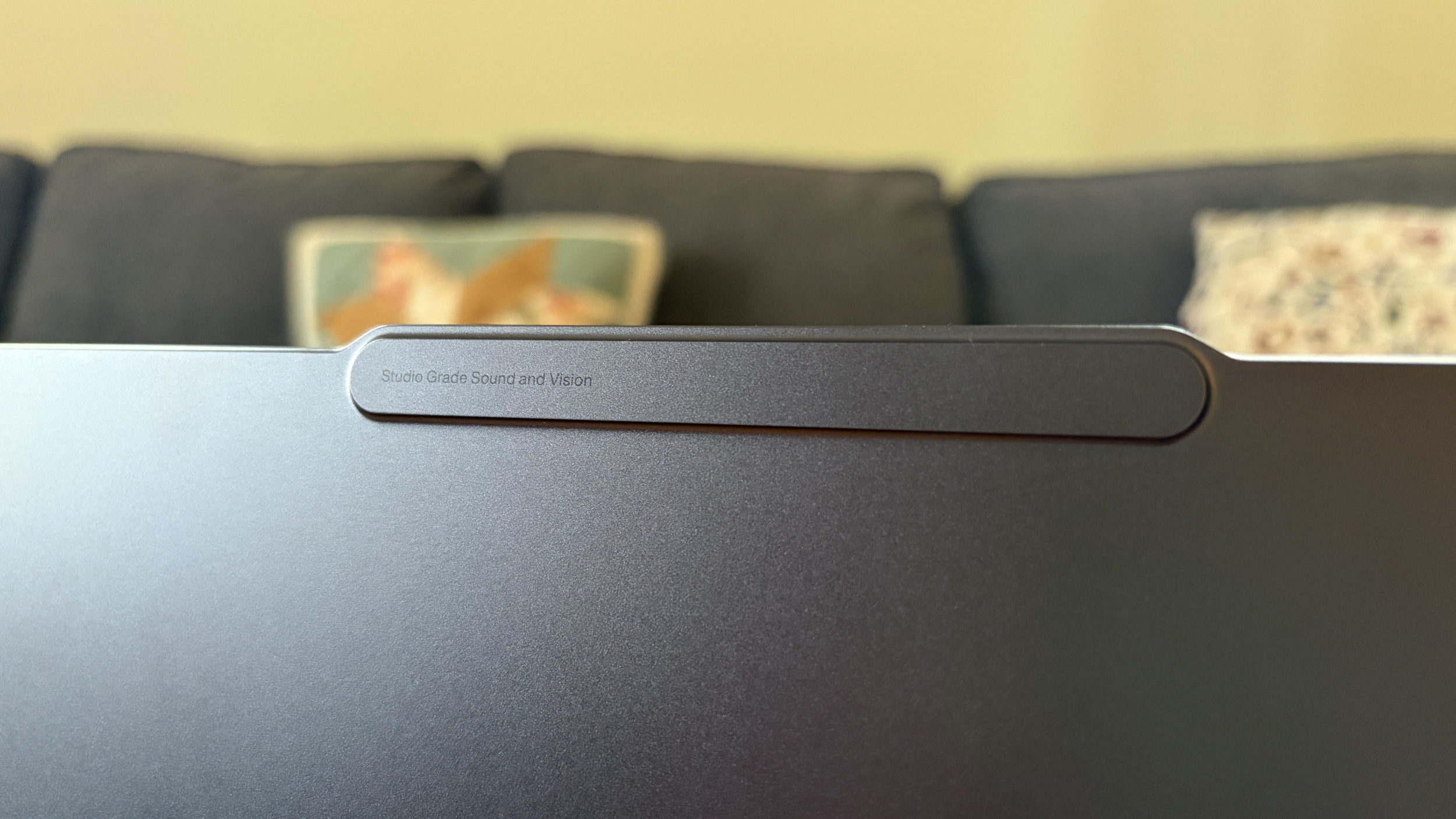 Close-up of a dark gray laptop or monitor edge with a slim, rounded bar; a softly blurred sofa and cushions create a calm indoor backdrop.