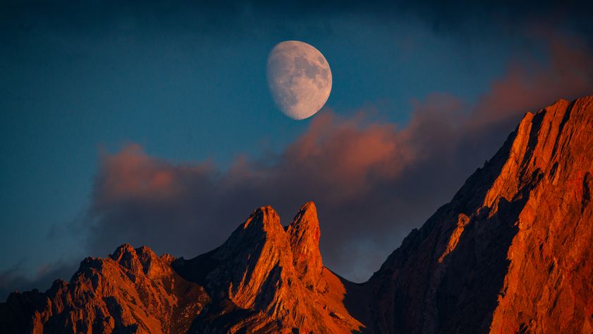 A near full moon is pictured in a blue sky above the jagged peaks of a mountain range. Both the mountains and the clouds below the moon are lit by the golden light of dusk.