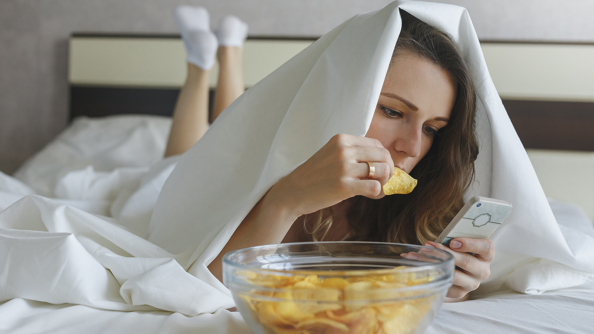 A woman lies on her bed beneath a duvet eating from a bowl of chips