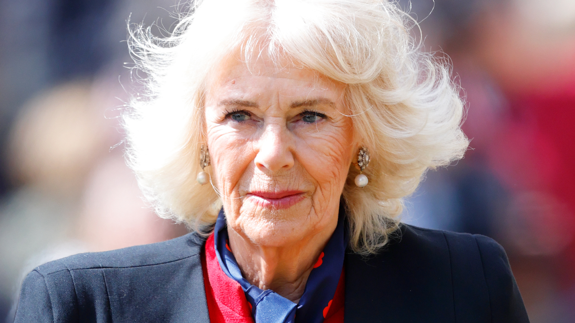 Queen Camilla wears a black blazer and a red dress and has blonde hair which is in an upheaval in the wind