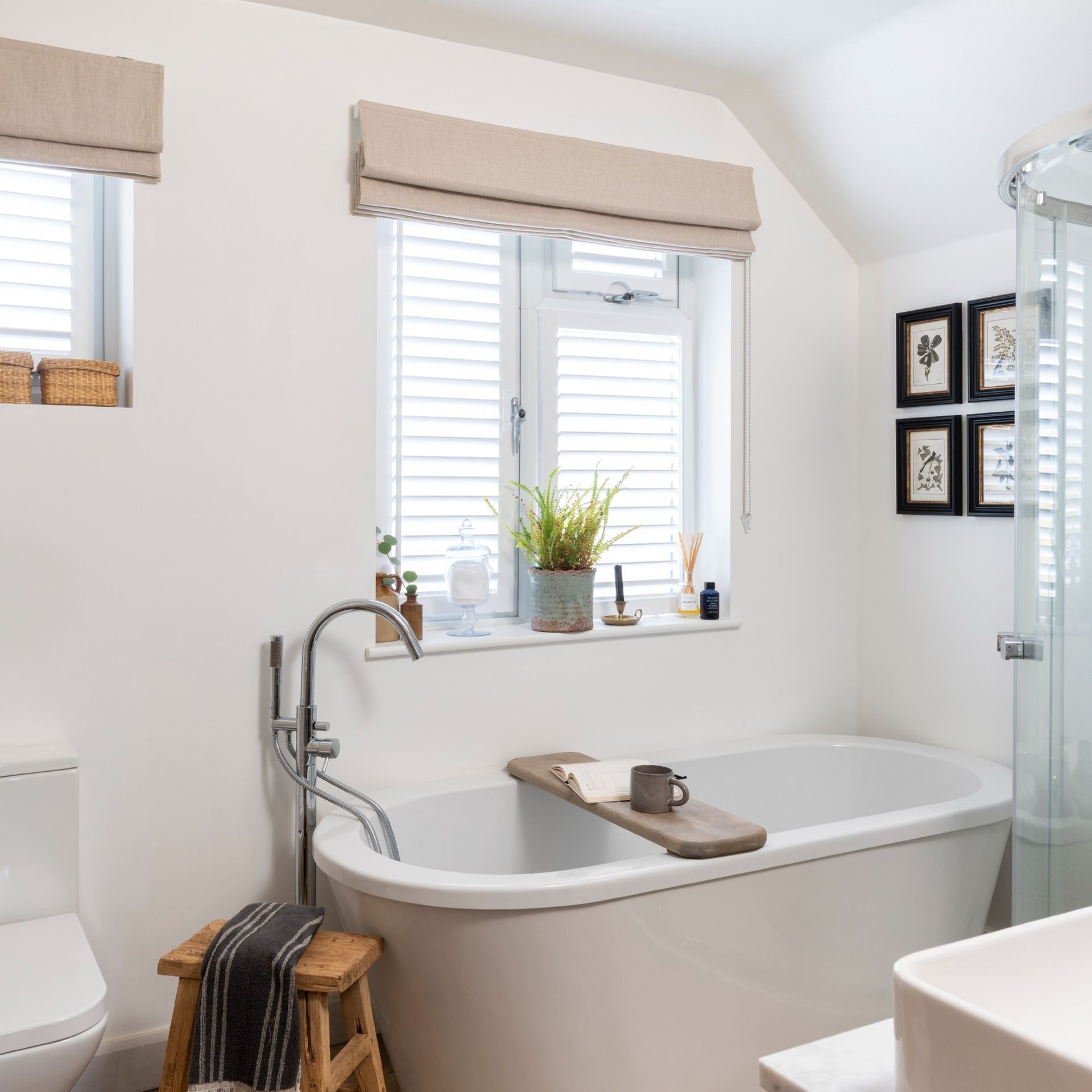 A white bathroom with a bathtub and windows  with both slatted blinds and Roman blinds