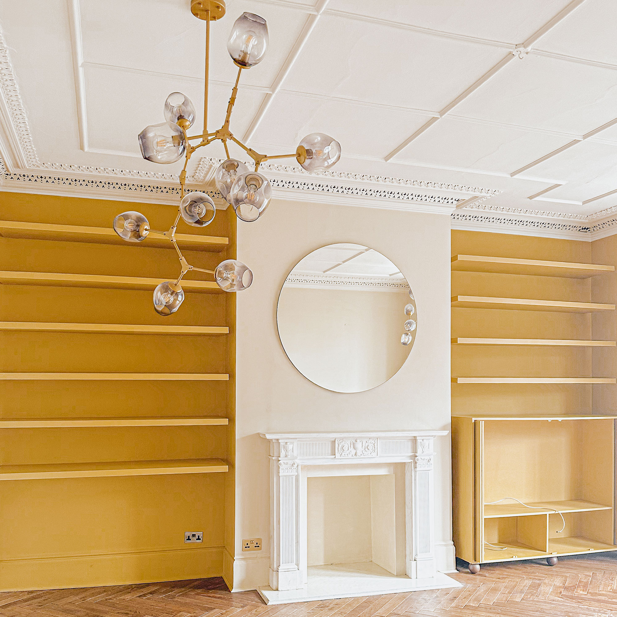Glass and brass sputnik style light in unfinished room with white panelled ceiling, fireplace, round mirror and yellow painted built-in bookshelves