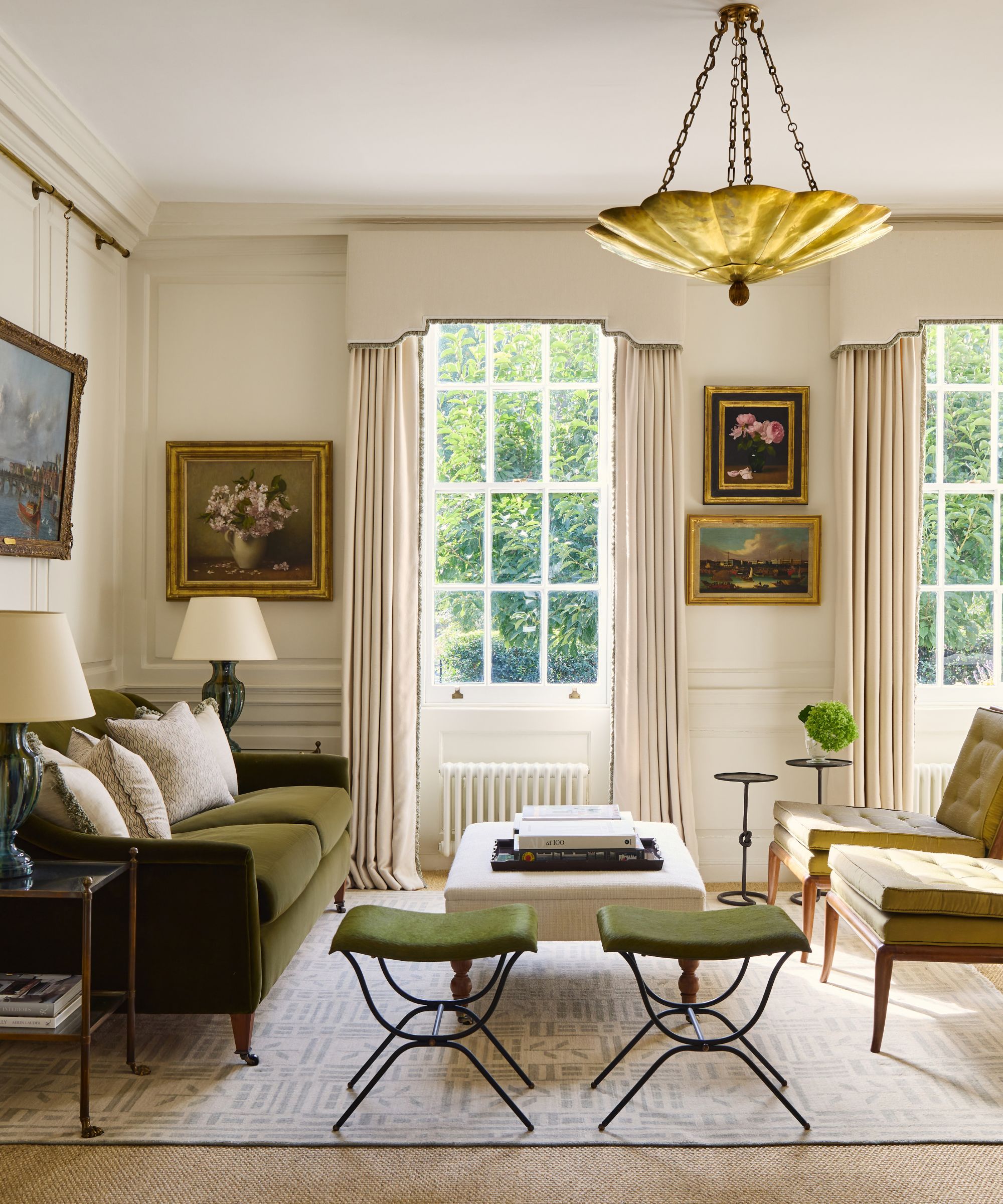 Living room with two long windows with pelmet curtains, a gold pendant light, two green wrought iron stools, a green velvet sofa, and two beige chairs, on top of a patterned rug.