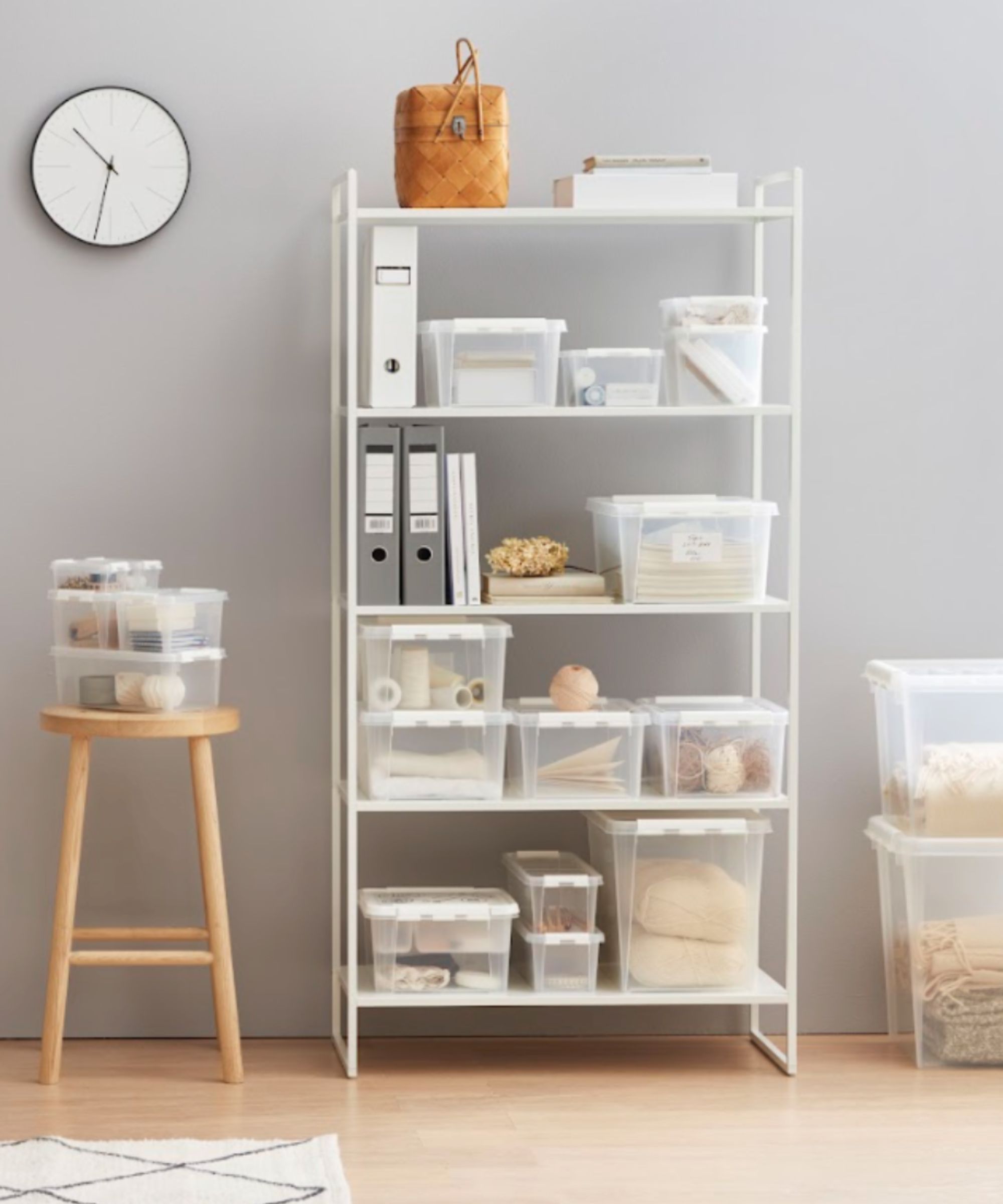 Organised home office corner with a white shelving unit and clear plastic storage boxes, neutral decor, a wooden stool, and a minimalist wall clock.