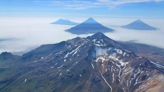 An aerial oblique photo of the volcanoes in the Islands of Four Mountains, Alaska, taken in July 2014. In the center is the summit of Mount Tana. Behind Tana are (left to right) Herbert, Cleveland and Carlisle Volcanoes.