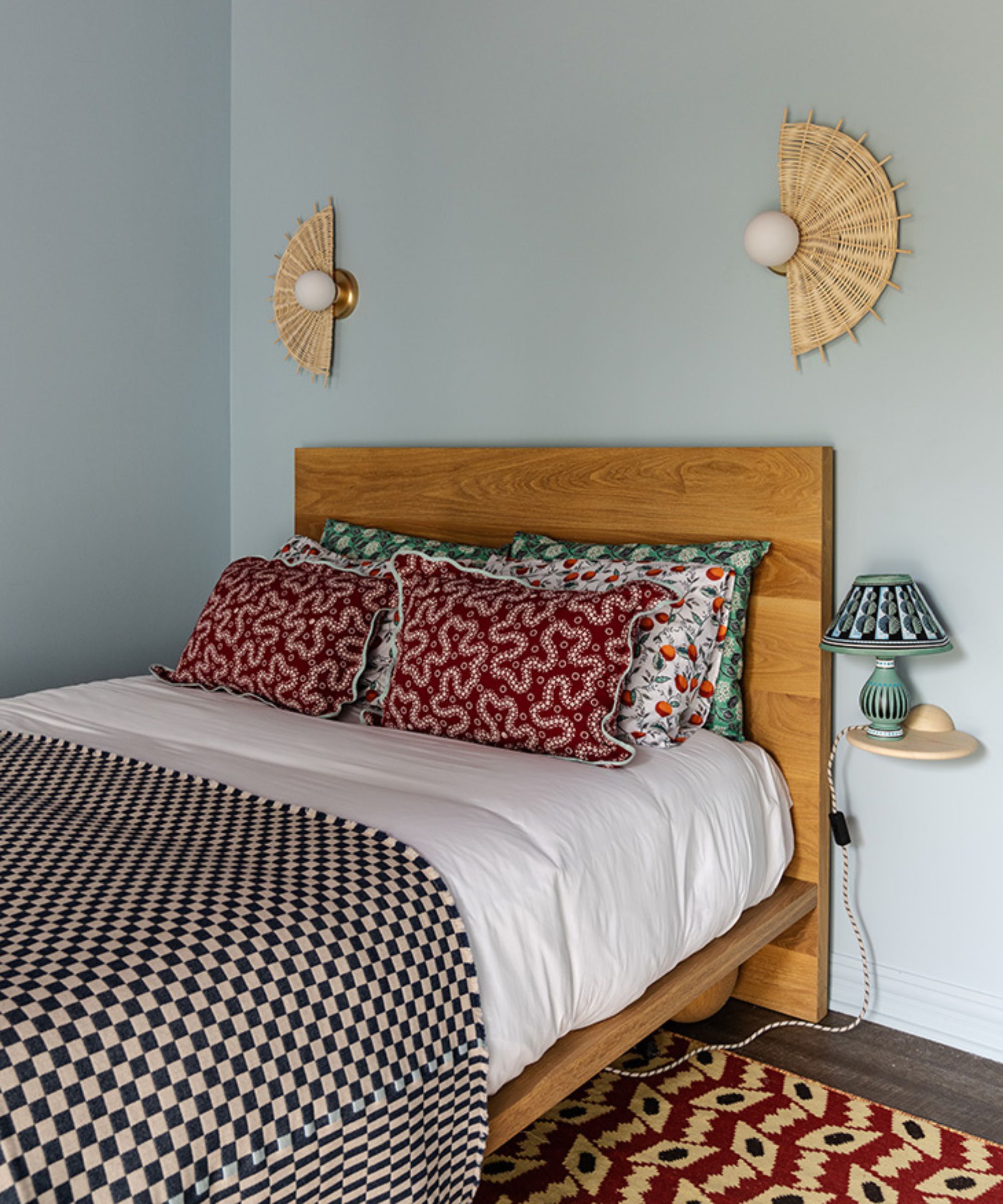 Light blue painted bedroom, wooden bed with colorful pillows, white bedding, black and white check throw. Two rattan wall lights above bed, patterned red and black rug. Small bedside table with lamp