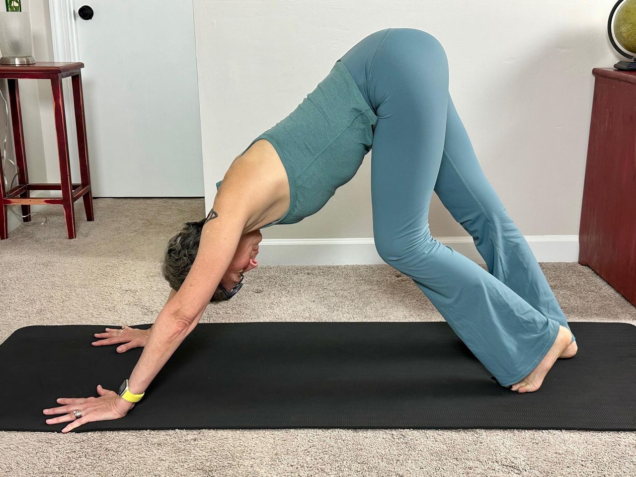 woman wearing blue vest and leggings performing a downward facing dog yoga pose on a black yoga mat in a living room