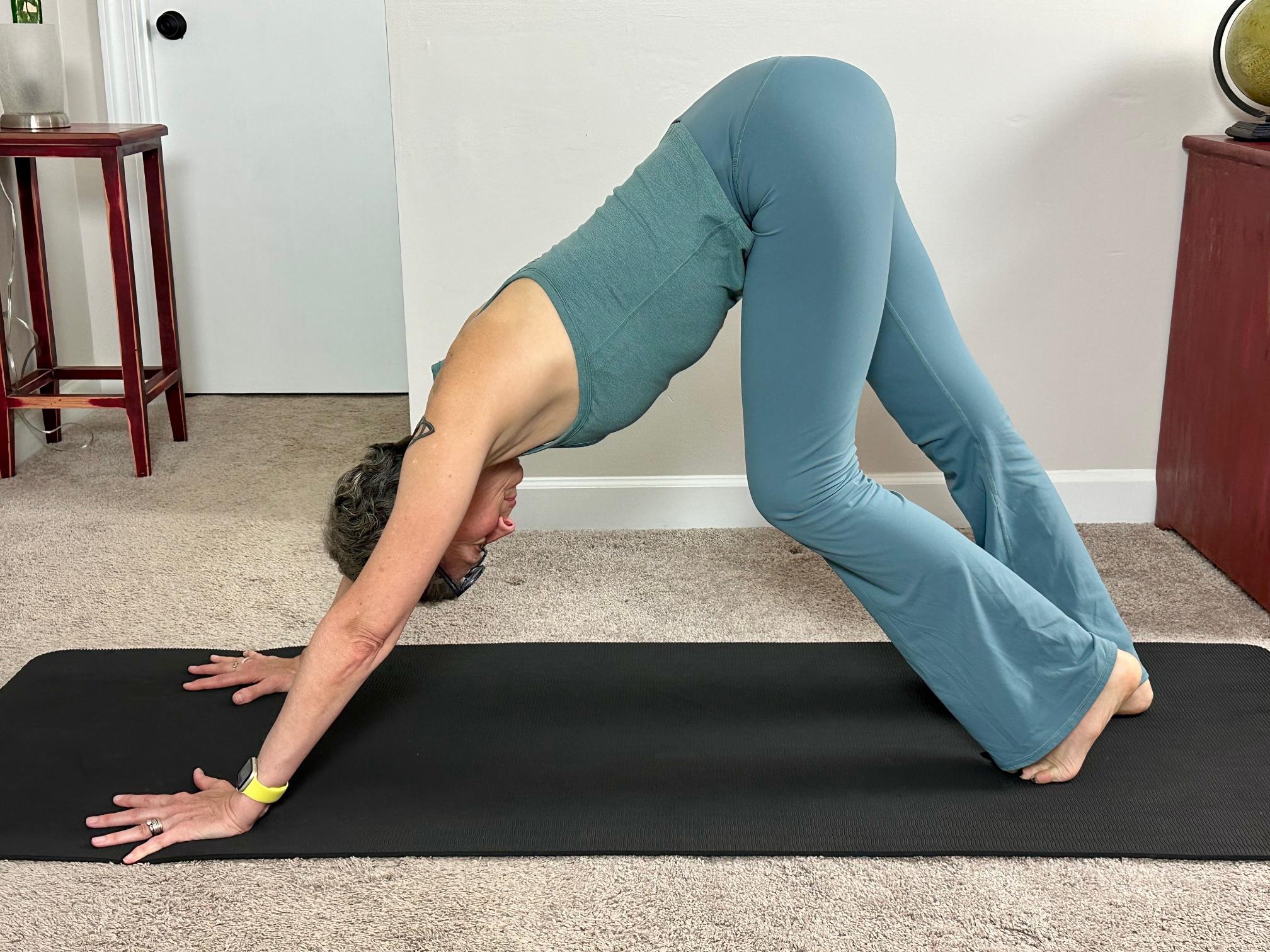 woman wearing blue vest and leggings performing a downward facing dog yoga pose on a black yoga mat in a living room