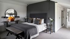 A white bedroom with grey feature wall, with a large bed with grey headboard, white bedding, a grey blanket, and grey and brown cushions in the center. At the foot of the bed is a leather bench. To the left of the bed is a wooden sidetable with a large orange glass vase, two matching lamps and a large circular mirror above, and to either side of the bed are wooden side tables with flowers. The floor is light grey carpet, and to the right of the room, white cabinets are seen, while the white door to the bedroom is open.