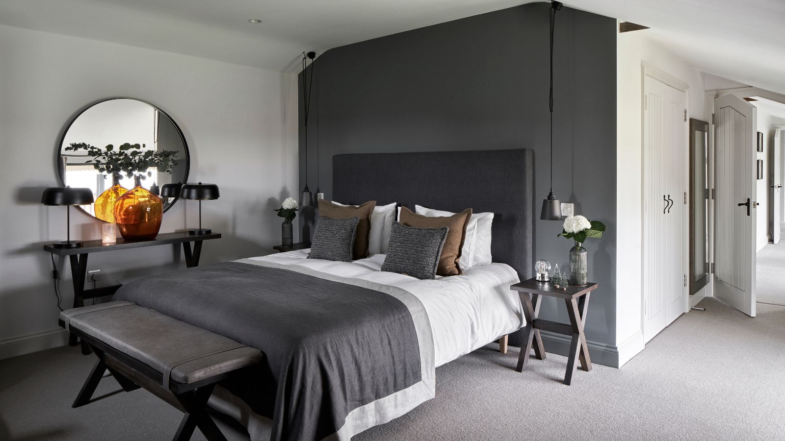 A white bedroom with grey feature wall, with a large bed with grey headboard, white bedding, a grey blanket, and grey and brown cushions in the center. At the foot of the bed is a leather bench. To the left of the bed is a wooden sidetable with a large orange glass vase, two matching lamps and a large circular mirror above, and to either side of the bed are wooden side tables with flowers. The floor is light grey carpet, and to the right of the room, white cabinets are seen, while the white door to the bedroom is open.