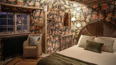 Moody cottage bedroom with floral wallpaper on the walls and low ceilings. It has a large pink bloom oa n black background design, the Divine Savages Forbidden Bloom Black wallpaper, with wicker chair and cozy bed with rounded natural headboard. The window shows dusk outsid and the mood is wintery