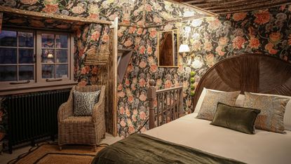 Moody cottage bedroom with floral wallpaper on the walls and low ceilings. It has a large pink bloom oa n black background design, the Divine Savages Forbidden Bloom Black wallpaper, with wicker chair and cozy bed with rounded natural headboard. The window shows dusk outsid and the mood is wintery