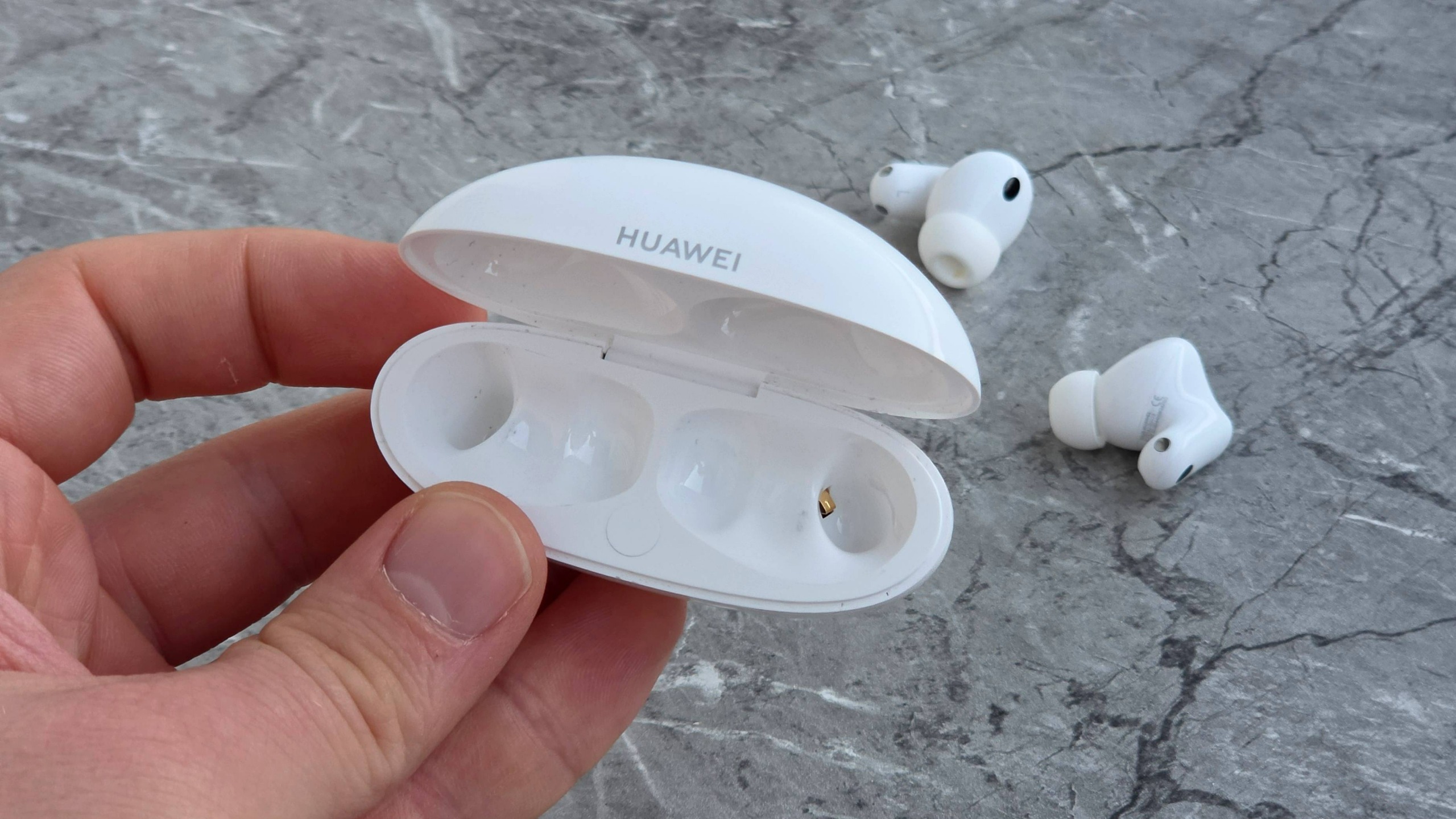 The Huawei FreeBuds Neo case in a man's hand, in front of the buds on a marbled surface.
