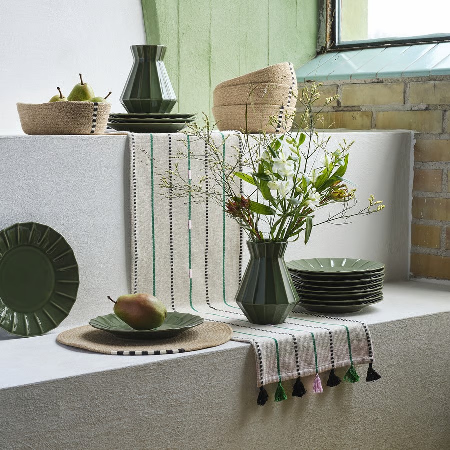 A collection of jute baskets and placemats mixed in with an assortment of green plates and vases. There is a white, embroidered table runner in the middle. Everything is arranged across two white steps.