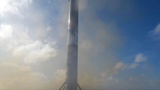 the first stage of a rocket stands vertical against a blue sky with white clouds after landing on an ocean-based droneship