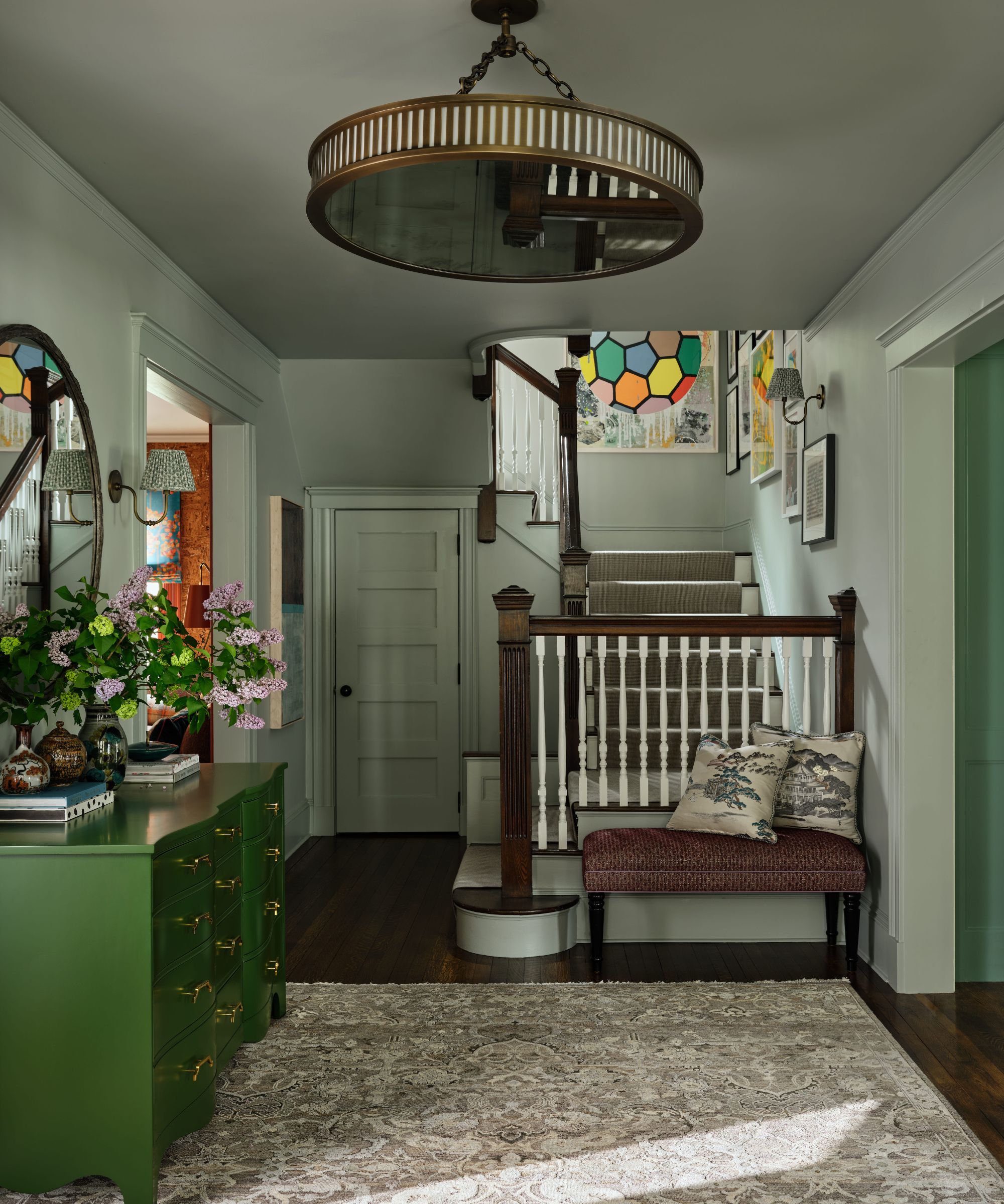 Light gray entryway with a green dresser, a wooden staircase with a striped runner, and a large round brass ceiling fixture.