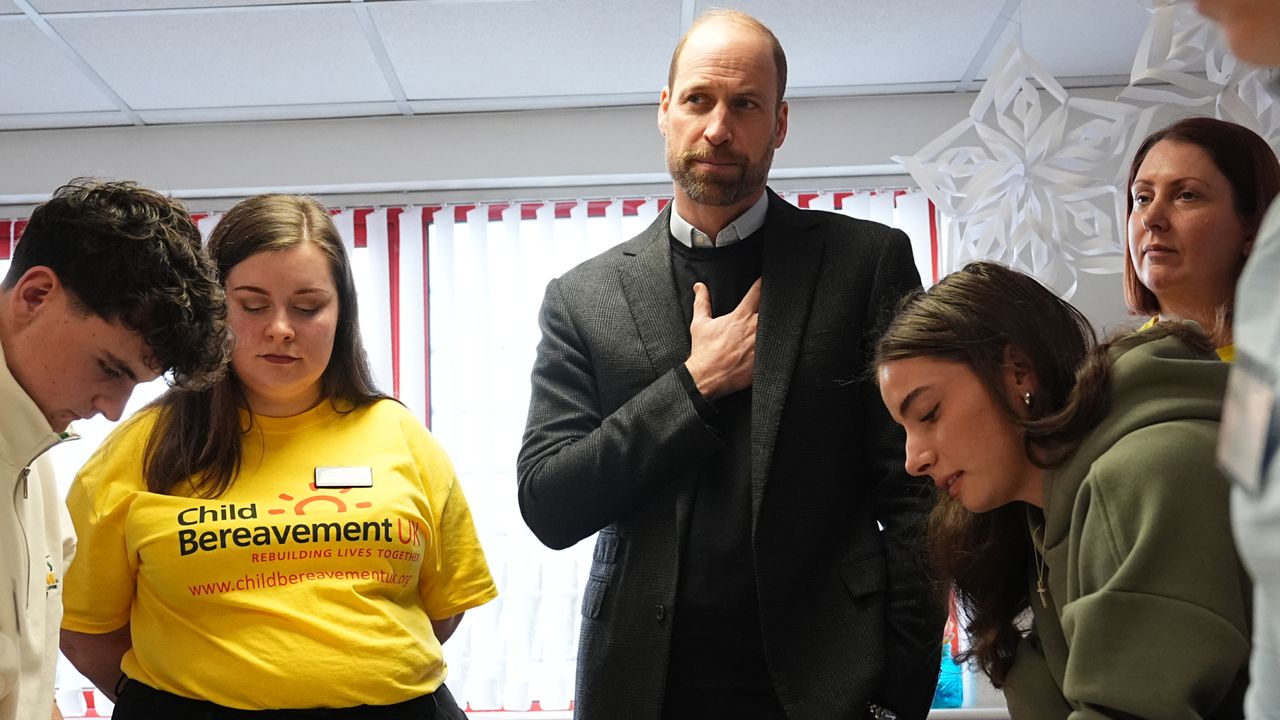 Prince William wearing a gray suit jacket putting his hand on his chest surrounded by a group of teenagers including a woman wearing a yellow Child Bereavement UK shirt