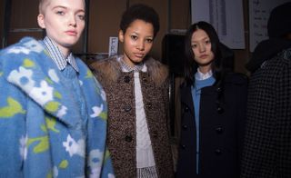 Three models, one in a pale blue fur coat with floral design, one in a brown duffel coat over a cream wool sweater, and one in a black coat over a blue sweater
