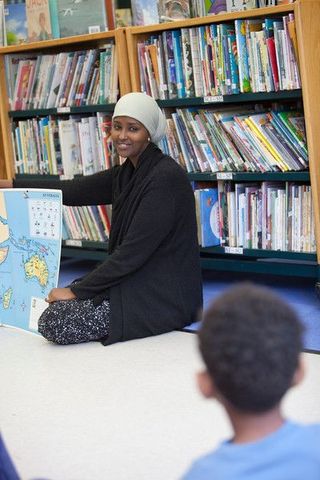 Fadumo Dayib on the floor in a school library with bookshelves behind her, addressing school children
