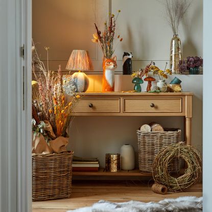 A hallway with a light wood console table decorated with a table lamp and dried flowers displayed in animal and mushroom-shaped vases and wicker baskets